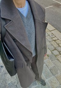 Brun ullfrakk, grå strikkegenser med V-hals, svart skulderveske, mørke bukser og svarte joggesko. Hette-design og synlige knapper.