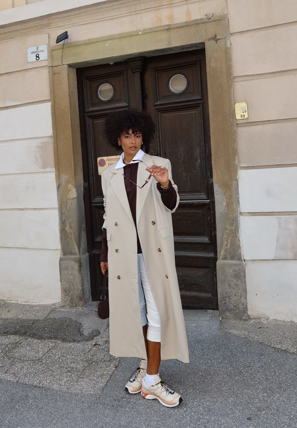 Beige doppelt knöpfter Trenchcoat, brauner Rollkragenpullover, weiße 7/8-Hosen und beige Sneakers mit bunten Akzenten; hält eine Sonnenbrille.