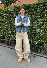 Farmerdzseki szürke pulóver felett, bézs bő nadrág és barna csizma. A háttérben buja zöld növényzet és egy fémkerítés látható.