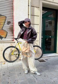 Smeđa sjajna skraćena jakna, prugasta košulja, bež torba s remenom, široke svijetle hlače, srebrne tenisice, stoji pokraj žutog bicikla.