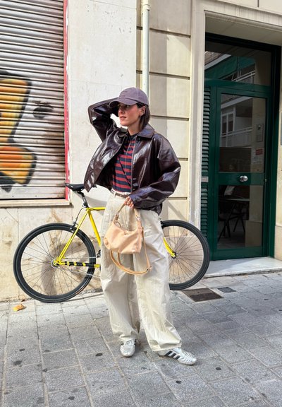 Chaqueta corta marrón brillante, camisa de rayas, bolsa beige con una correa, pantalones ligeros holgados, zapatillasplateadas, de pie junto a una bicicleta amarilla.
