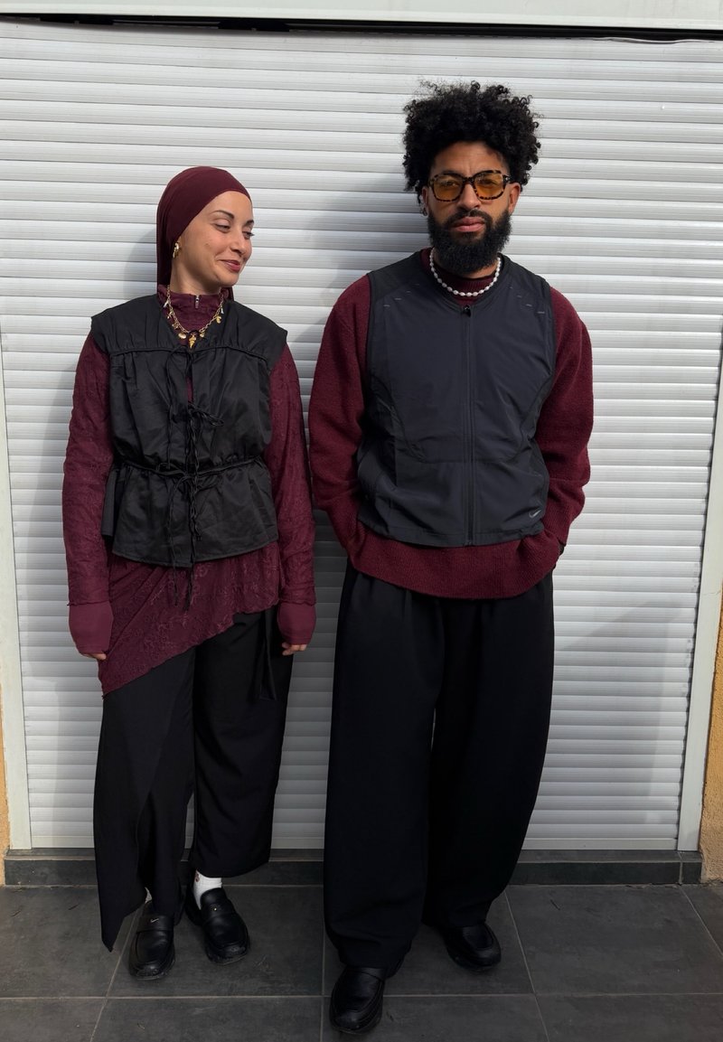 Deux personnes se tiennent côte à côte devant une porte roulante blanche ; toutes deux portent des hauts bordeaux, des gilets noirs, des pantalons noirs amples et des chaussures noires.