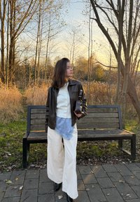 Giacca di pelle marrone, maglione bianco con bottoni, sciarpa blu, pantaloni bianchi a gamba larga, stivali neri, in piedi in un ambiente naturale con alberi e prato.