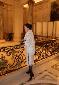 Blondetørklæde i hvidt over en satin kjole med knælængde og elegant flow. Sorte hæle fuldender looket foran et udsmykket gelænder.