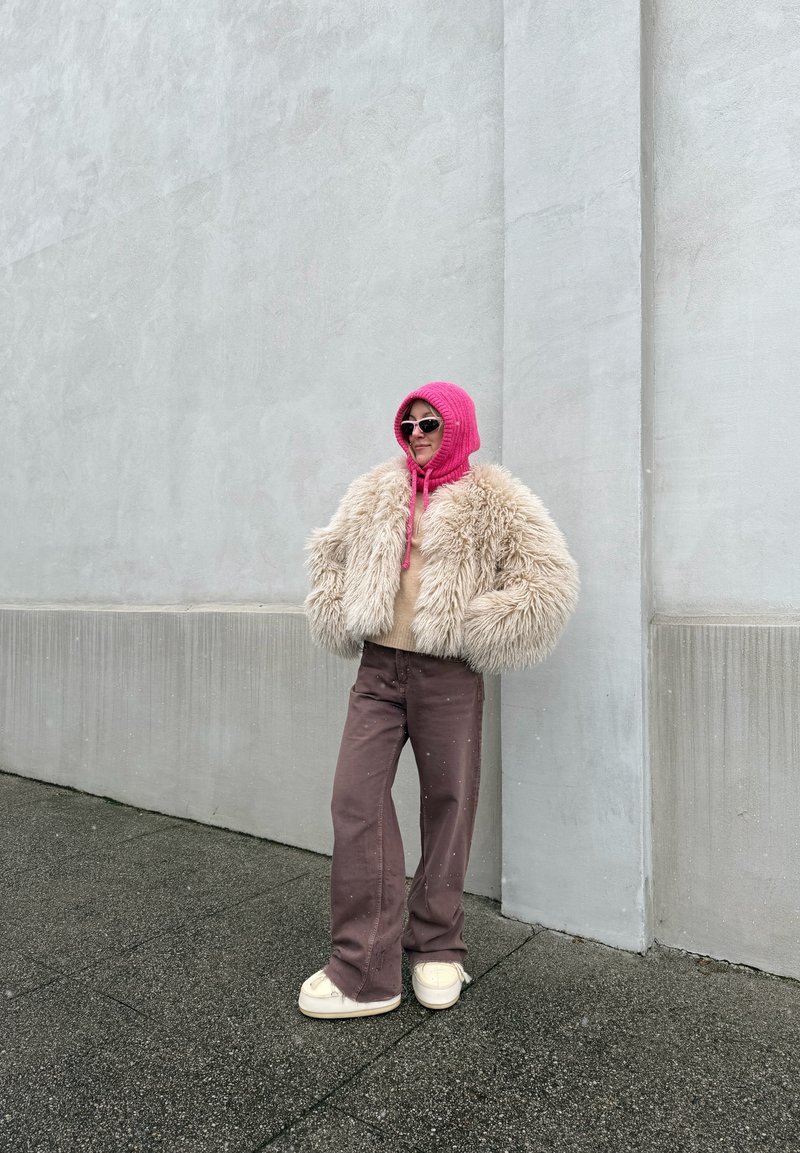Veste en fausse fourrure crème avec manches oversize, sweat à capuche en tricot rose, pantalon évasé marron et chaussures à plateforme crème, posés devant un mur gris.
