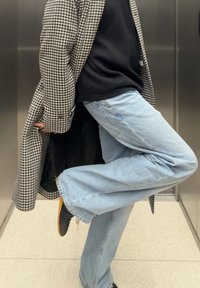 Houndstooth-kåpe i svart og hvitt, lagd over en svart topp. Lyseblå utsvingte jeans og svarte sko med brun såle. Innendørs miljø.