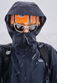 Crna vodootporna jakna s visokim ovratnikom i zatvaranjem na patentni zatvarač, uparena s narančastom pletenom kapom i bijelim sunčanim naočalama, nasuprot snježnoj pozadini.