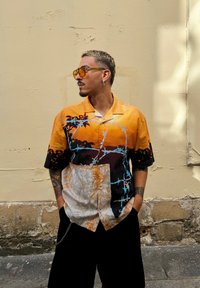 Μπλουζάκι κοντό μανίκι, πορτοκαλί και μαύρο με μοτίβα που παρουσιάζουν αφηρημένα σχέδια και μπλε λεπτομέρειες, σε χαλαρή γραμμή και με κουμπωτή μπροστινή πλευρά.