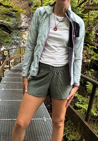 Lagani mint jakna s uzorkom i crnim detaljima, uparenih s maslinasto zelenim šortsevima. Vanjsko okruženje s drvenim ogradom i lišćem.