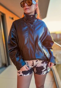 Černá bunda z umělé kůže s vysokým límcem, nadměrným střihem a patentními detaily, doplněná béžovými šortkami s černým vzorem připomínajícím skotu.