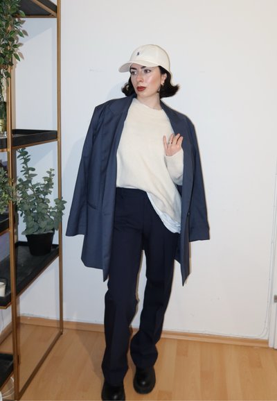 Blazer azul marino de gran tamaño, suéter crema, pantalones azul marino y botas negras. La gorra de béisbol blanca añade contraste. Diseño simple y minimalista con tonos neutros.