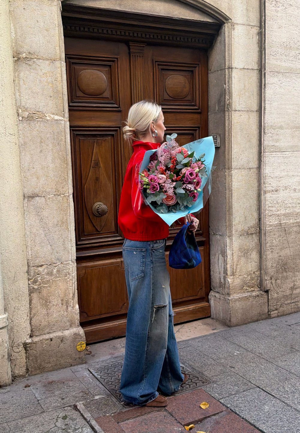 Femme aux cheveux blonds portant une veste rouge et un jean large, tenant un bouquet de fleurs et un sac bleu, debout devant une grande porte en bois.