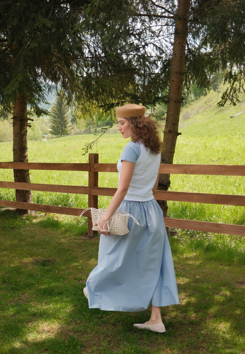 Femme en jupe bleu clair et chapeau beige marchant sur l'herbe près d'une clôture en bois et d'arbres dans une prairie verte ensoleillée.