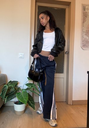 Giacca bomber nera, canotta bianca corta, pantaloni Adidas blu navy con strisce bianche, borsa nera e scarpe da ginnastica beige. Ambiente interno con piante.