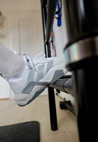 Osoba w białych i szarych butach sportowych opiera stopę na pedale czarnego metalowego roweru treningowego wewnątrz budynku.