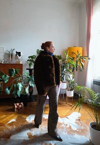 Femme debout à l'intérieur près d'une fenêtre, portant un pantalon imprimé léopard et une veste en fourrure marron foncé, entourée de plusieurs plantes vertes en pot et de meubles en bois.