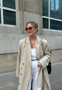 Beige dupla soros blézer nagy gombokkal, fehér top, magas derekú fehér nadrág, barna öv, mintás napszemüveg, fekete kézitáska.