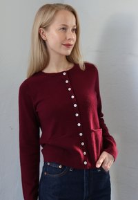 Bordo kardigan s dugim rukavima, okruglim izrezom, prednjim kopčanjem na dugmad i dva prednja džepa, izrađen od meke tkanine glatke teksture.