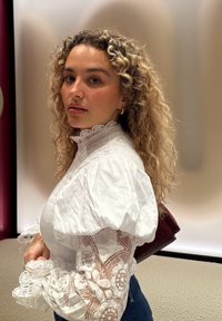 Camicia bianca con maniche a sbuffo, dettagli in pizzo sulle braccia, abbinata a jeans scuri, su uno sfondo con luce soffusa.