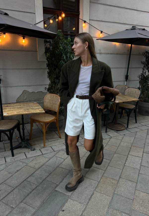 Lang olijfgroene jas, witte shorts, lichte top en hoge bruine laarzen. Buitensetting met lichtsnoeren en café tafels. Neutraal kleurenpalet.