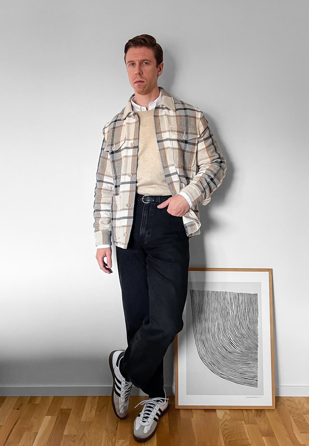 Homme portant une veste à carreaux beige, un pull crème, un jean noir et des baskets blanches, appuyé contre un mur blanc à côté d'une œuvre d'art en noir et blanc encadrée.