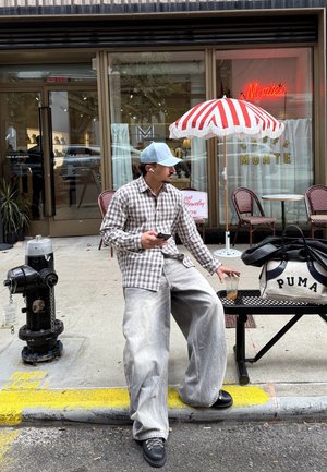 Camisa de cuadros abotonada en marrón y beige, vaqueros holgados gris claro, zapatos negros, gorra azul, sosteniendo un teléfono inteligente y una bebida helada. Bolsa negra con texto blanco.