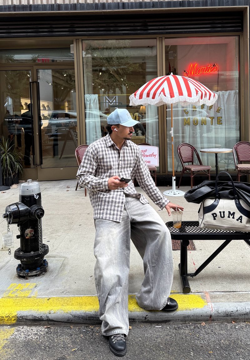 Koszula na guziki w brązowo-beżową kratę, obszerne jasnoszare dżinsy, czarne buty, niebieska czapka, trzyma smartfon i zimny napój. Czarna torba z białym napisem.