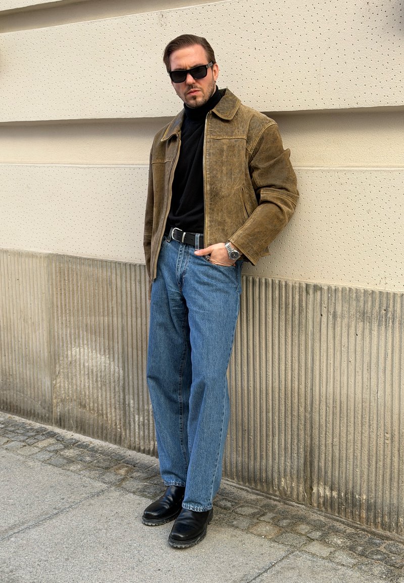 Homme portant des lunettes de soleil, une veste en daim marron, un col roulé noir, un jean bleu et des bottes noires, appuyé contre un mur texturé beige en extérieur.
