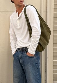 Weißes langärmeliges Henley-Shirt mit Knopfleiste, blaue Jeans aus Denim und grüne Wildleder-Schultertasche, stehend vor einer neutralen Wand.