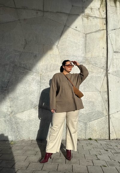 Jersey marrón oversize, pantalones de pierna ancha crema, botines burdeos y un bolso cruzado marrón contra una pared de concreto geométrica.