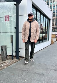 Μπεζ επενδυμένο μπουφάν, μαύρη κουκούλα, πράσινα παντελόνια, γκρι αθλητικά παπούτσια. Υπαίθριος χώρος με γυάλινο κτίριο, άνδρας με γυαλιά ηλίου και καπέλο.