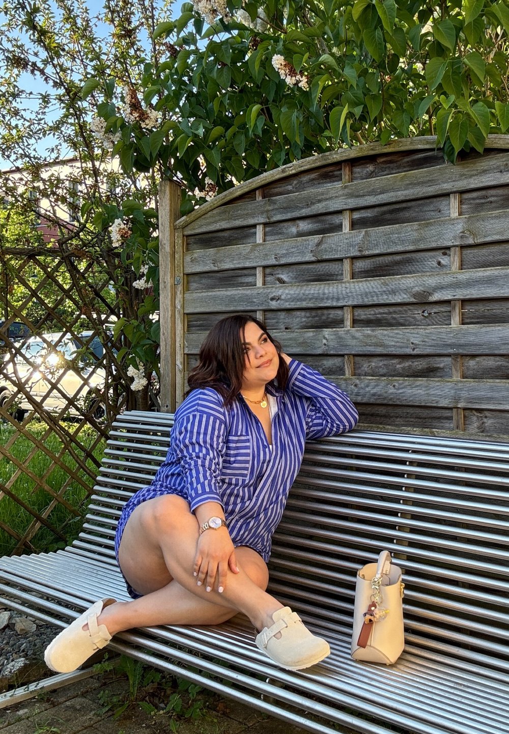 Camicia a righe blu e bianca, calzature chiare, seduto su una panchina metallica, con una borsa crema accanto, sullo sfondo di verde.