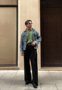 Man wearing black wide-leg trousers, a green shirt, a denim jacket, and sunglasses, standing on a pavement next to a beige wall and a dark door.
