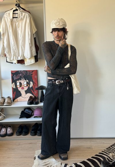 Persona con tatuajes lleva un suéter oscuro transparente, pantalones anchos negros, gorra clara y bolsa de mano, de pie en una habitación con zapatos, gorras y camisetas colgadas.