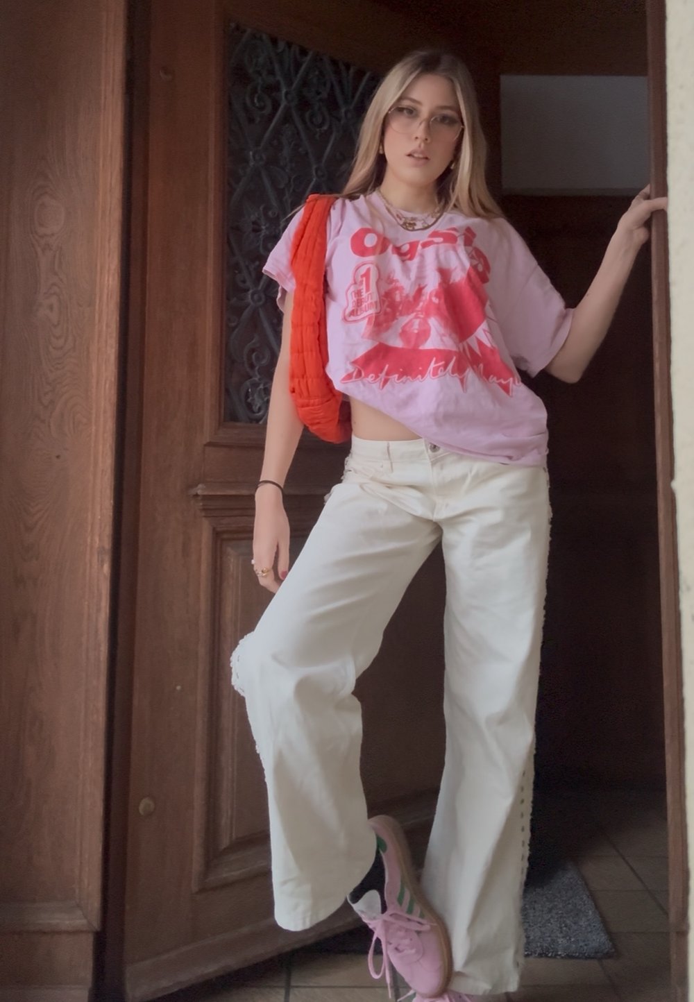 Νεαρή γυναίκα με γυαλιά ακουμπάει στην πόρτα, φοράει ροζ μπλουζάκι με στάμπα, λευκό παντελόνι φαρδύ, ροζ αθλητικά παπούτσια και κόκκινη τσάντα στον ώμο.