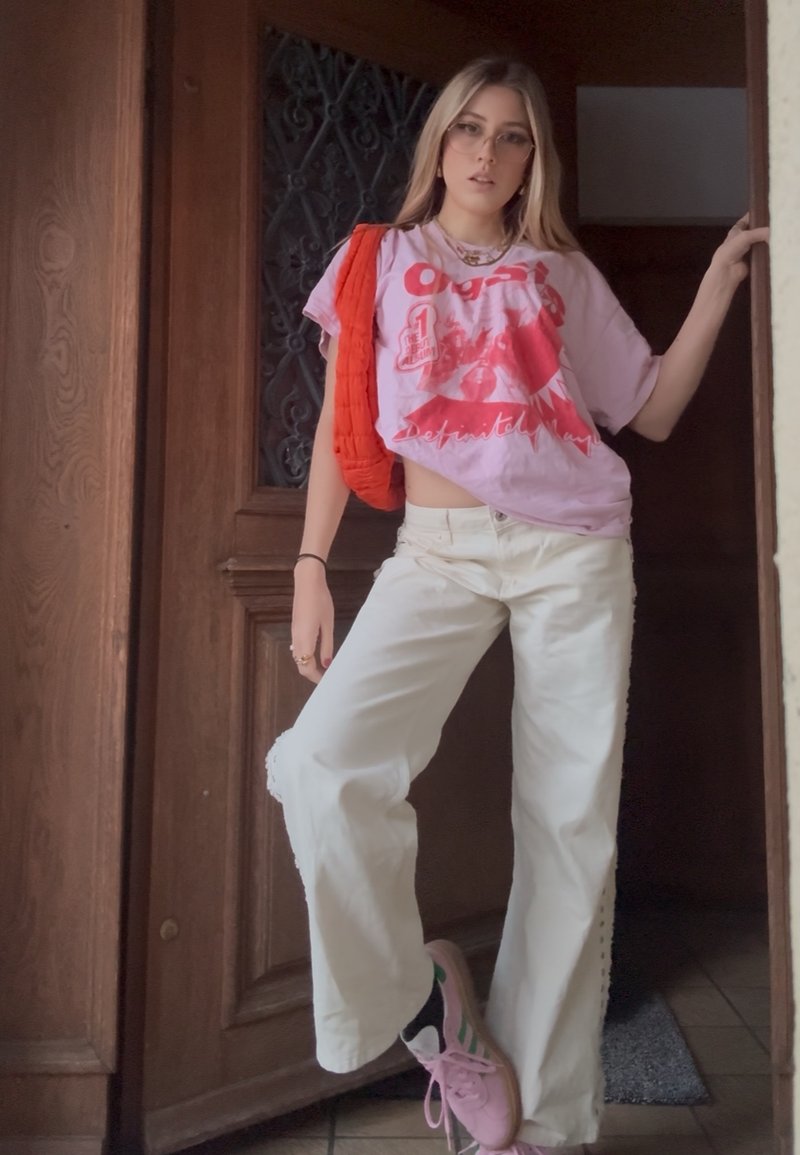 Junge Frau mit Brille lehnt an einem Türrahmen, trägt ein pinkes T-Shirt mit Grafik, weiße weite Hosen, pinke Turnschuhe und eine rote Schultertasche.