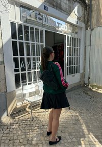 Black backpack with multiple compartments, worn over a green sweatshirt with pink stripes, paired with a black skirt and flat shoes, standing near a shop.