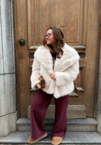 Femme aux longs cheveux bruns portant des lunettes, une veste blanche duveteuse, un pantalon grenat et des chaussures beige, debout devant une porte en bois tenant une pochette marron.