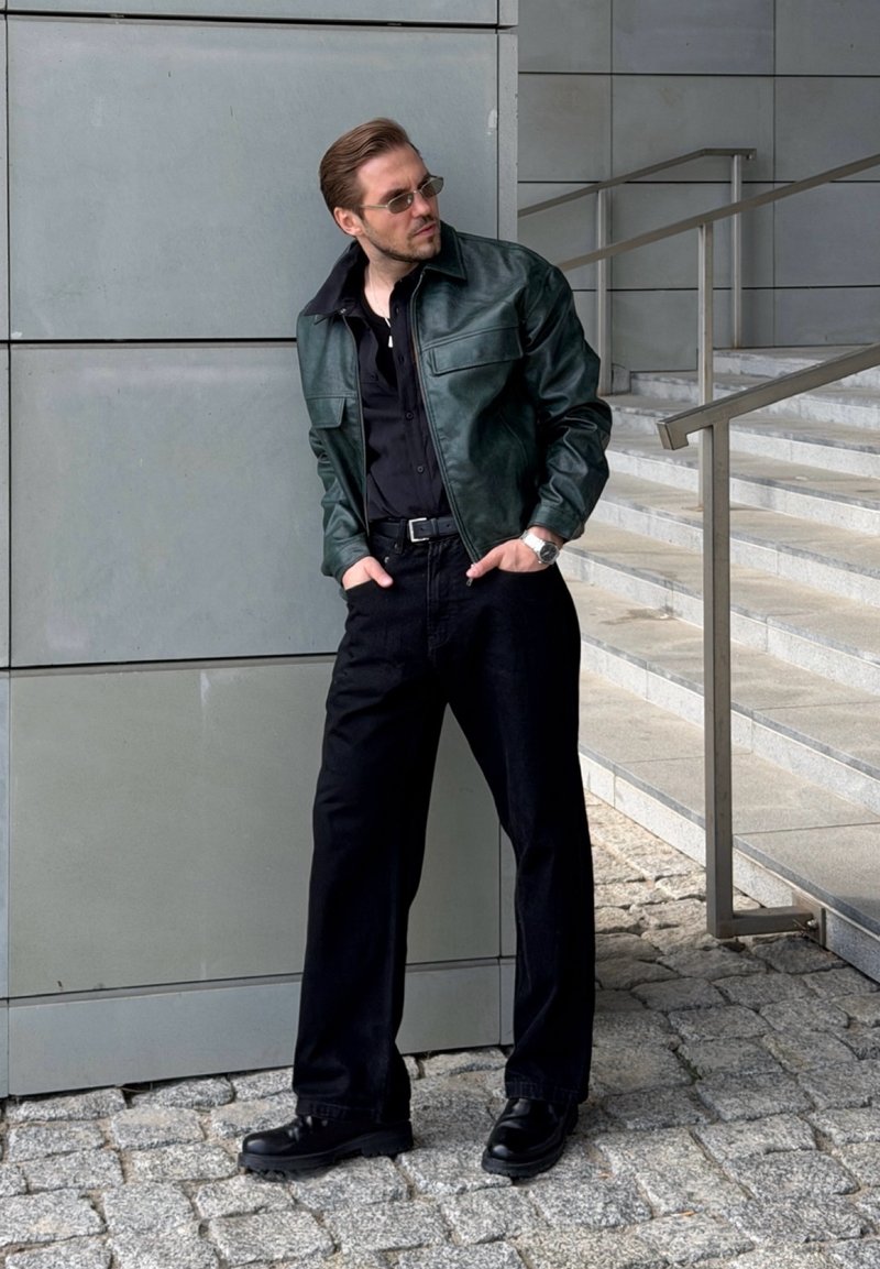 Homme portant une veste en cuir vert foncé, une chemise noire, un pantalon noir et des lunettes de soleil, debout près d'un mur carrelé gris et d'un escalier avec une rampe métallique.