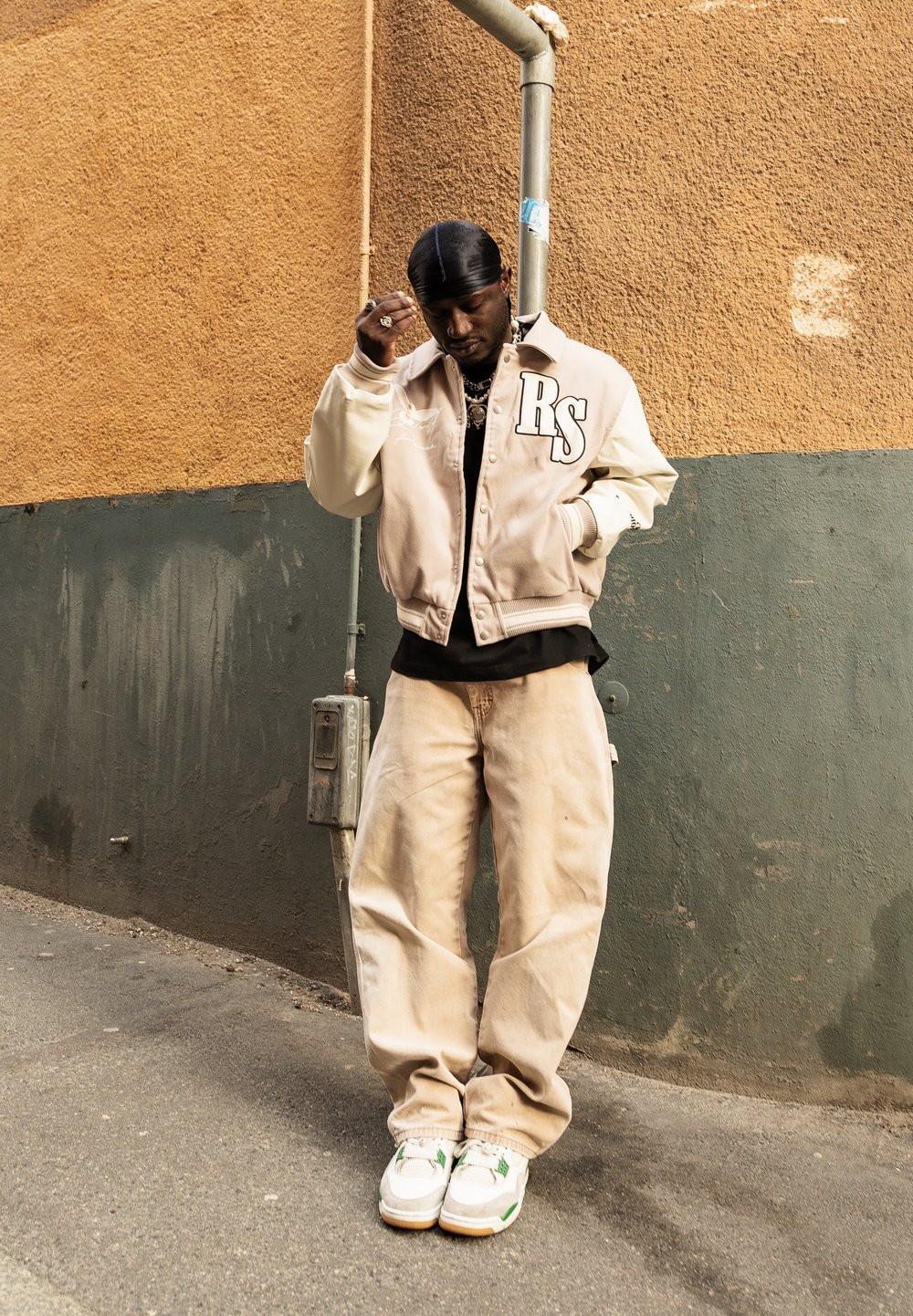Homme portant une veste universitaire beige avec les lettres « RS », un pantalon beige, des baskets blanches et un durag noir, debout devant un mur bicolore vert et orange.