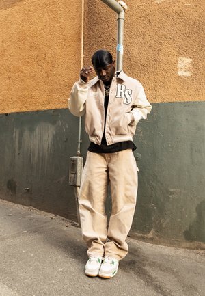 Hombre con chaqueta universitaria beige con "RS," pantalones color canela, zapatillas blancas y un durag negro, de pie frente a una pared bicolor verde y naranja.