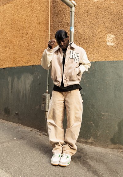 Hombre con chaqueta universitaria beige con "RS," pantalones color canela, zapatillas blancas y un durag negro, de pie frente a una pared bicolor verde y naranja.