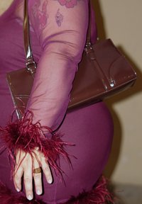 Naiste käsi läbipaistvas tumepunasest varrukast sulekanuga, ülakehal tätoveering, hoides pruuni nahast käekotti, sõrmes kuldsõrmus.