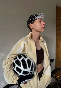Beige zip-up jacket with white accents, black sports bra, black cycling cap featuring a bicycle print, white bicycle helmet, grey wall, and a bike in the background.