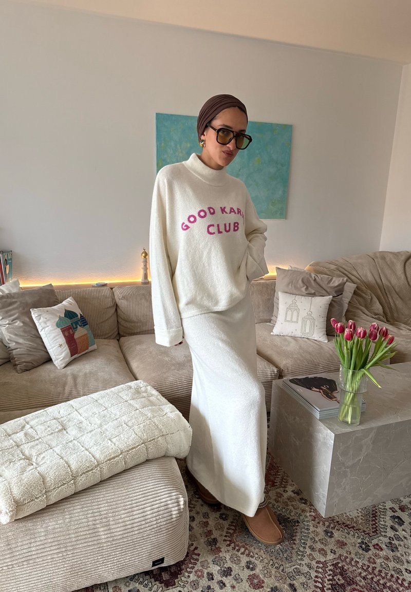 Femme portant un pull blanc "Good Karma Club" et une jupe, debout dans un salon avec un canapé beige, des tulipes dans un vase et une œuvre murale.