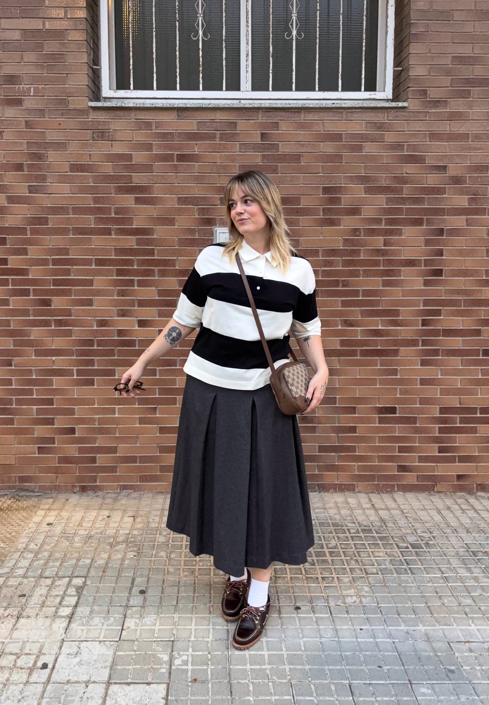 Polo a righe bianco e nero, gonna plissettata grigia, borsa a tracolla marrone e scarpe marroni con calzini bianchi contro un muro di mattoni.