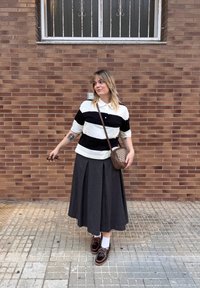 Polo a righe bianco e nero, gonna plissettata grigia, borsa a tracolla marrone e scarpe marroni con calzini bianchi contro un muro di mattoni.