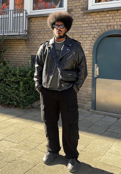 Hombre con peinado afro que lleva una chaqueta de cuero negra, pantalones cargo, zapatos grises, gafas de sol y una bolsa de bandolera, de pie en una acera soleada.