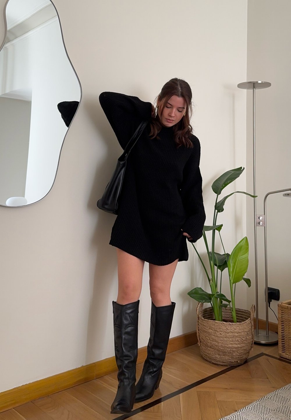 Vestido de malha preto com mangas longas, comprimento acima do joelho, combinado com botas altas pretas. Inclui uma bolsa de ombro e uma planta em vaso ao fundo.