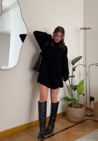 Vestido de malha preto com mangas longas, comprimento acima do joelho, combinado com botas altas pretas. Inclui uma bolsa de ombro e uma planta em vaso ao fundo.